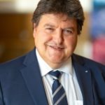 Close-up of a man smiling, wearing a blue suit with a striped tied