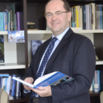 Foto Frans Vos - 1_1 Man in front of a bookcase holding a folder, smiling to the camera