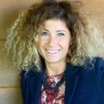 Joanna Wojewoda-Budka Woman in blue jacket and curly hair smiling in front of a wooden panel background